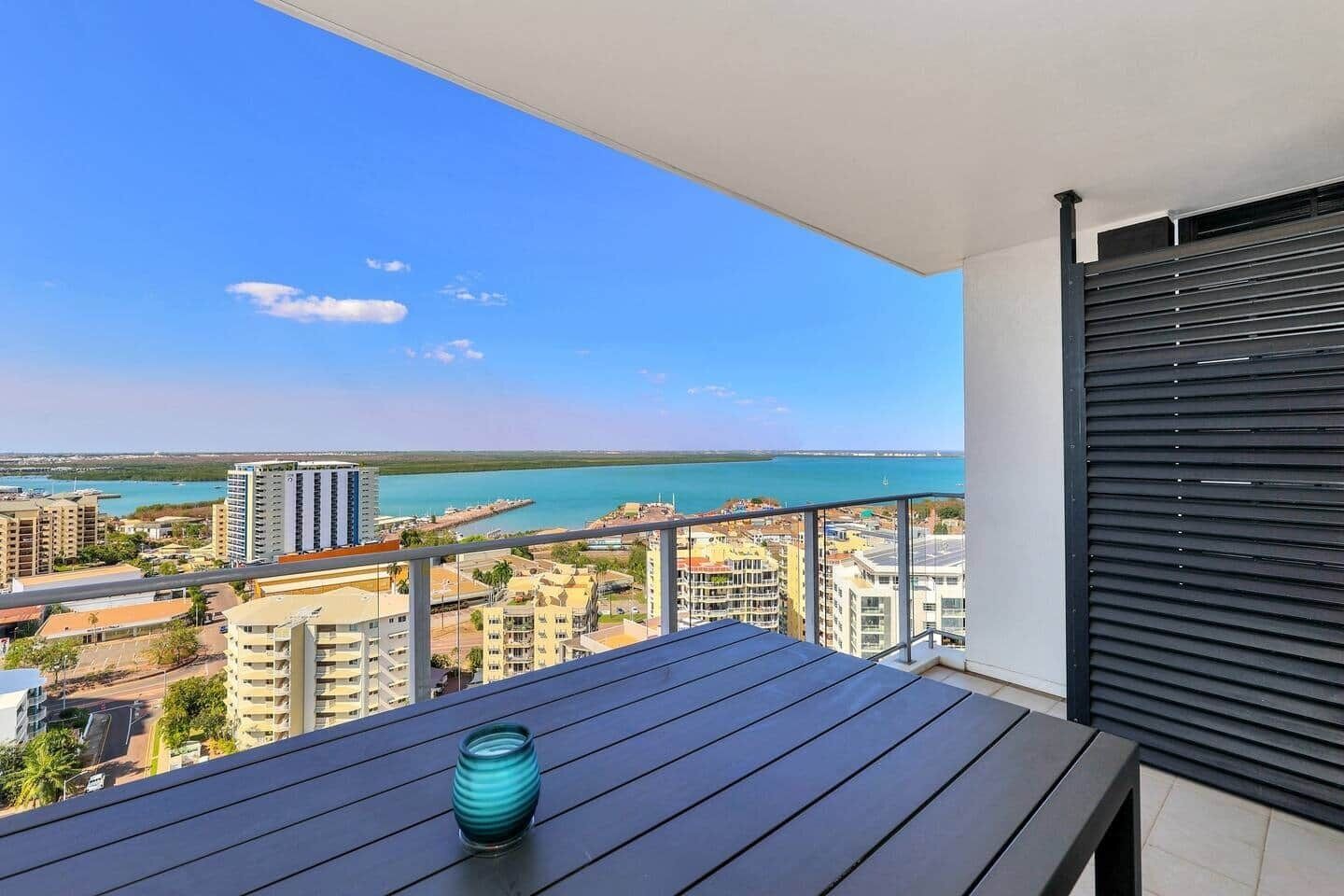 A balcony with a view of a coastal cityscape, a table with a vase, and a closed roller shutter.