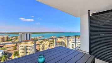 A balcony with a view of a coastal cityscape, a table with a vase, and a closed roller shutter.