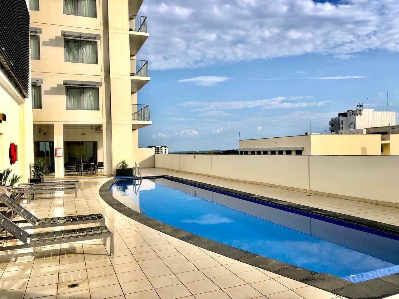 A swimming pool with lounge chairs, a building with balconies, and a clear sky.