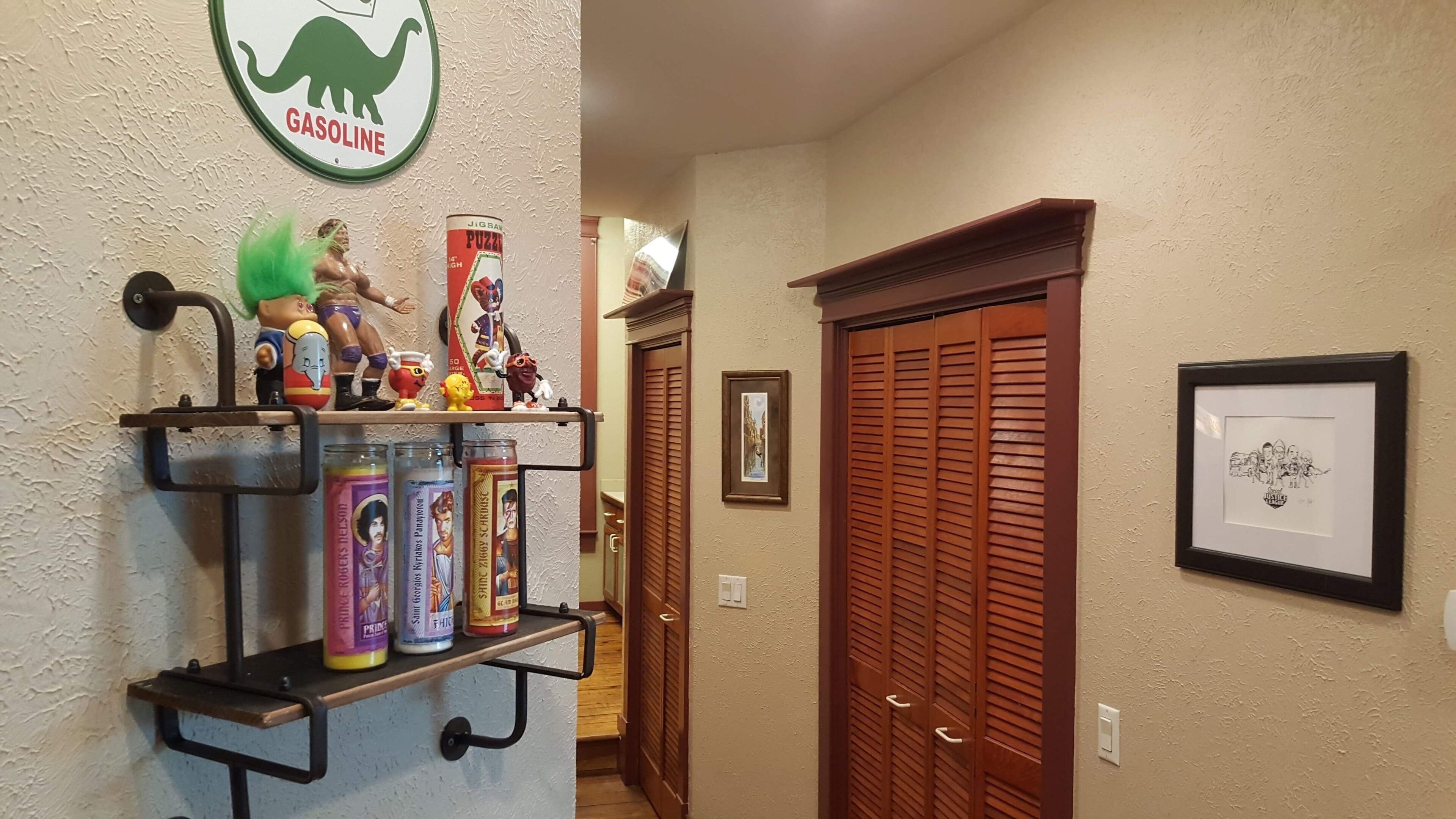 A hallway with a shelf displaying various items, including a dinosaur-themed sign, a puzzle box, and several cans. There are wooden doors and a framed picture on the wall.