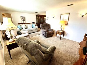 A living room with a sofa, armchair, and a small table with a lamp. There is a door leading to another room.