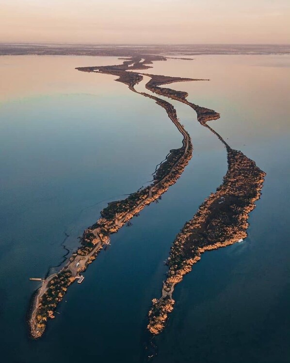 Vista aerea di una zona costiera con una lunga e stretta penisola che si protende nell'acqua, con una strada e alcuni edifici sparsi.
