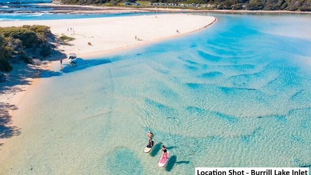 A beach with clear turquoise water, a person paddleboarding, and a sandy shore with buildings in the background. - Bring the Dog, Bring the Boat, Bring the Family (Burrill Lake)