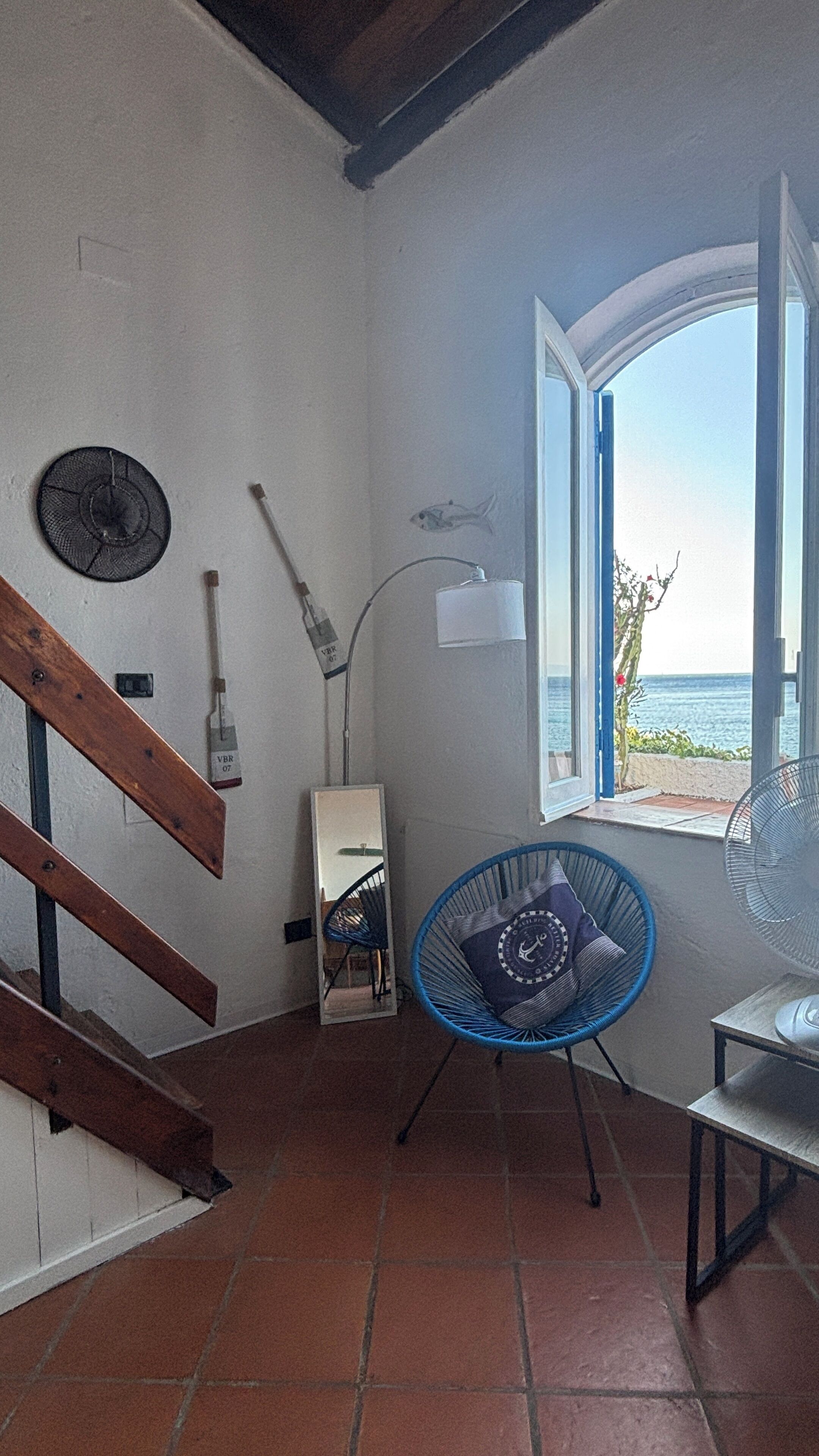 A room with a wooden staircase, a blue chair, a table, a window with a view of the sea, and a wall-mounted fan.