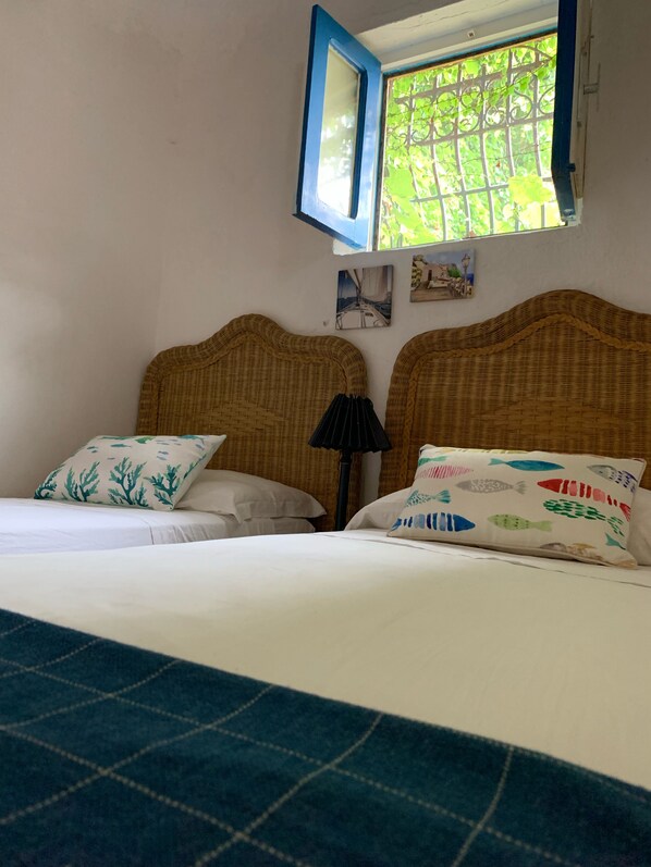 A hotel room with two beds, a window with a view of greenery, and a wall-mounted lamp.
