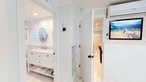 A bathroom with a white vanity, a mirror, a sink, and a toilet. There is a television mounted on the wall displaying a beach scene.