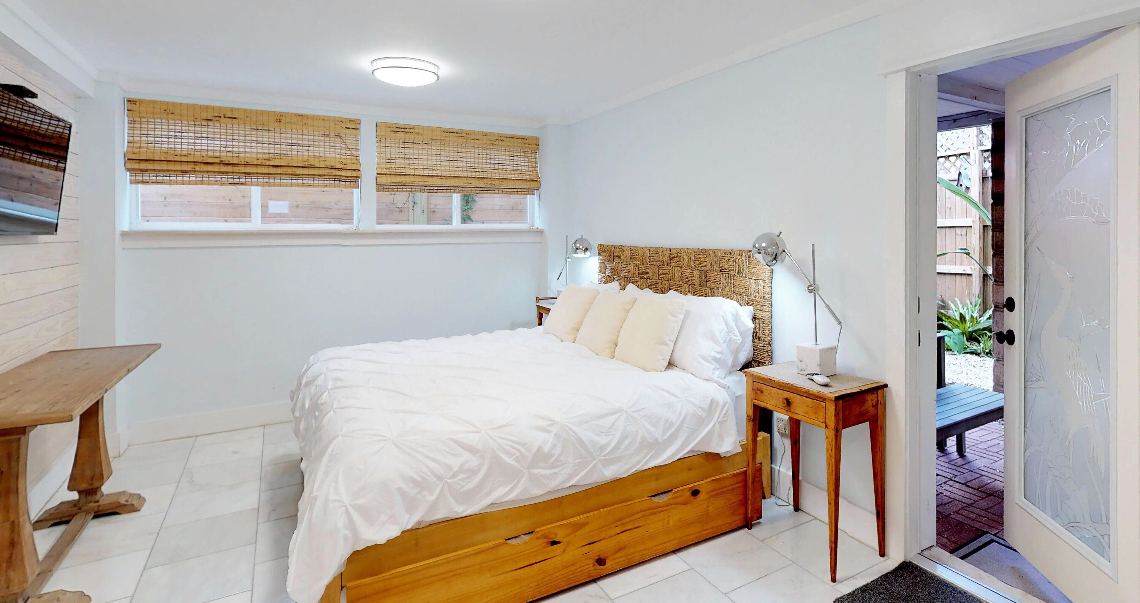 A bedroom with a wooden bed, white bedding, a bedside table with a lamp, and a window with bamboo blinds.