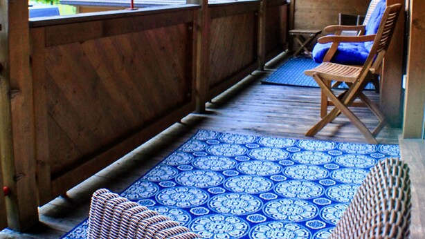 A wooden balcony with a patterned floor, two wooden chairs, and a view of a house and mountains.
