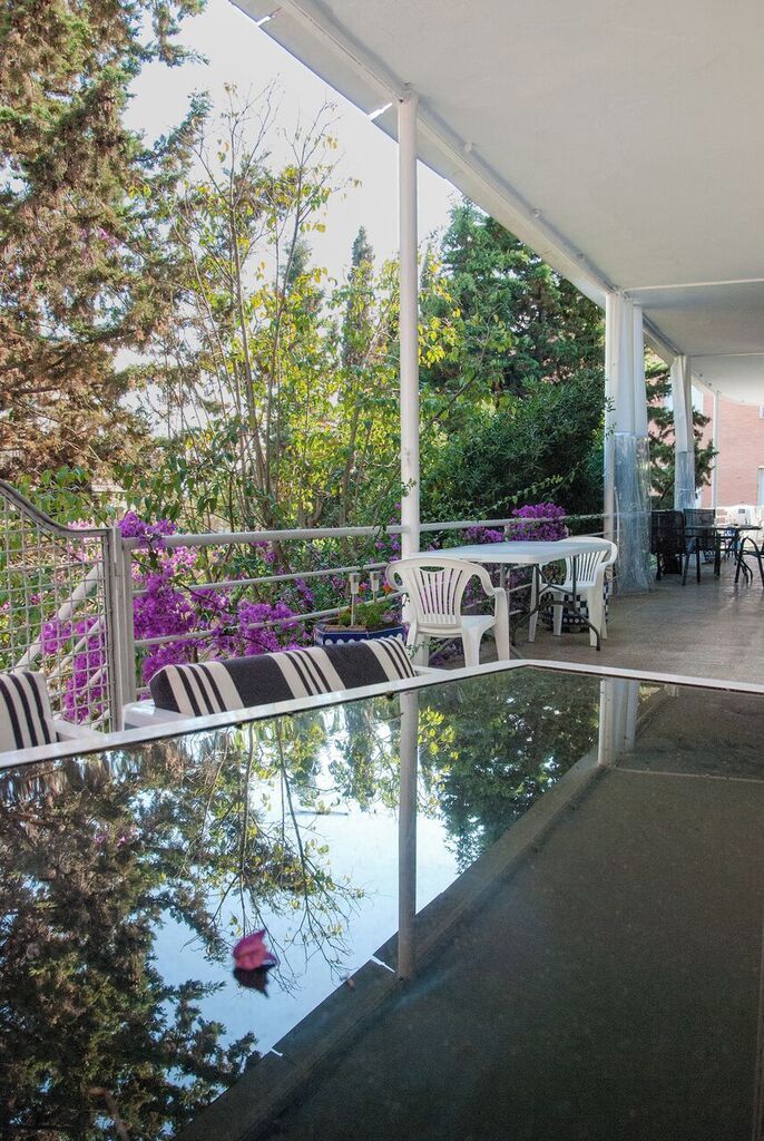 Un patio al aire libre con sillas y mesas blancas, rodeado de follaje y flores moradas.