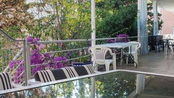 Un patio al aire libre con sillas y mesas blancas, rodeado de follaje y flores moradas.