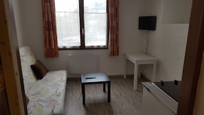 A small, sparsely furnished room with a sofa, a small table, a TV mounted on the wall, and a radiator. - STUDIO 17m², 5 BEDS (Les Orres)