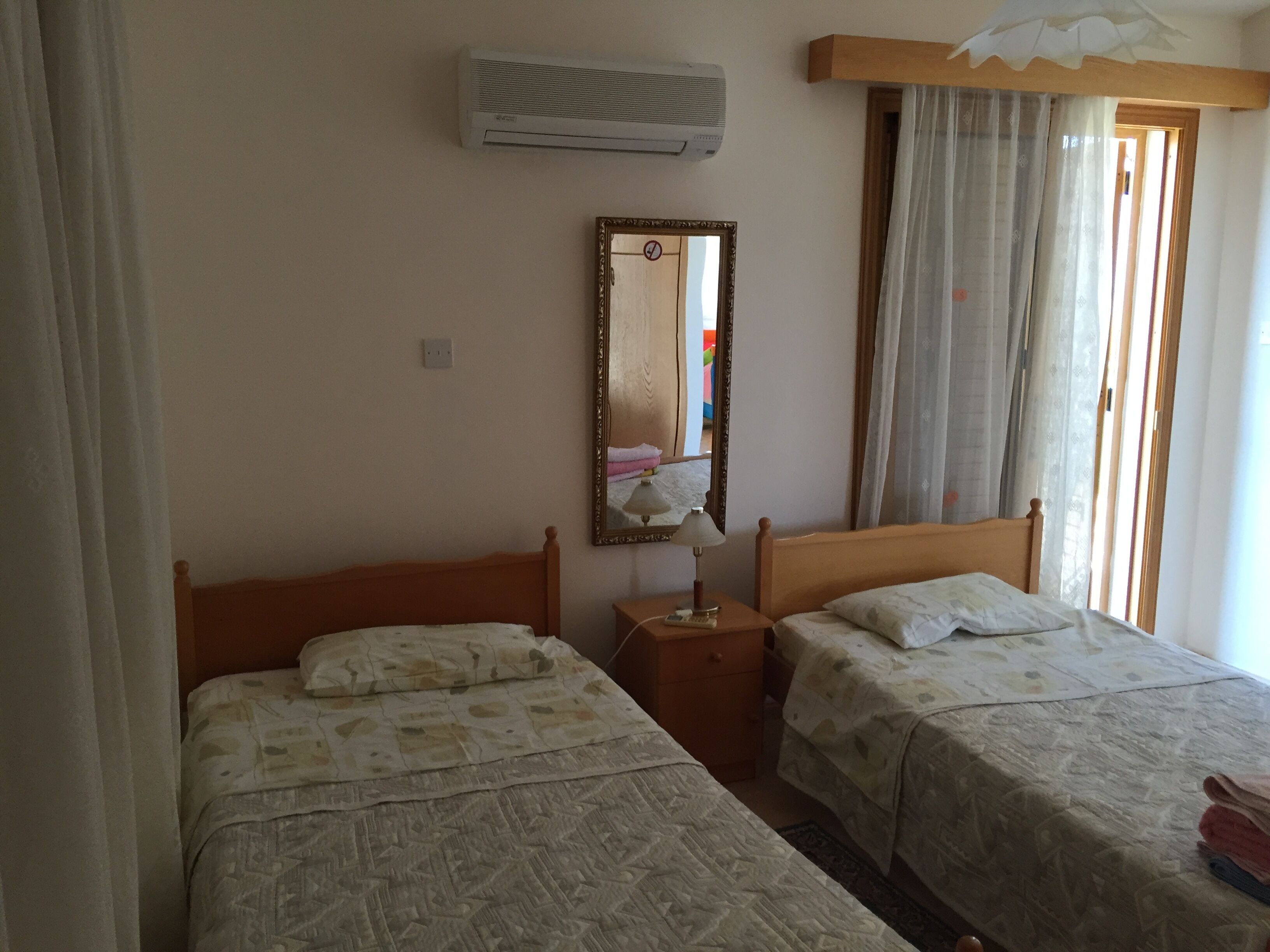 Une chambre avec deux lits simples, une table de chevet avec une lampe, un miroir, un appareil de climatisation et une fenêtre avec des rideaux.