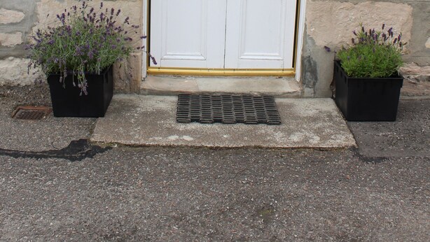 A stone building with a white door, two potted plants, and a sign above the door. - Beautiful cottage by the sea. (Nairn)