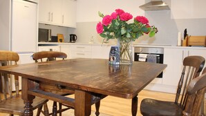 A wooden dining table with chairs, a vase with flowers, and kitchen appliances in the background. - Beautiful cottage by the sea. (Nairn)