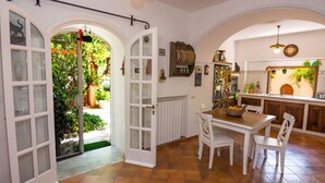 Une cuisine avec des meubles blancs, une table à manger et des chaises. Des portes ouvertes donnent accès à un jardin.