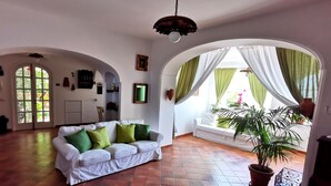 Un salon spacieux avec un canapé blanc, des coussins verts, une plante en pot et une grande fenêtre ornée de rideaux verts.