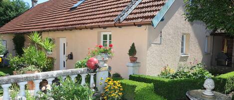Une maison avec un toit recouvert de tuiles rouges, un jardin où poussent différentes plantes et fleurs, et une fontaine.