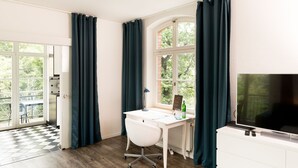 A room with a white desk, a chair, a TV, and a window with curtains. - LE Home II: 2-room loft w/ balcony+parking. Quiete, bright! (Plagwitz)