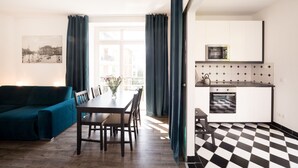 A modern living room with a sofa, dining table, and chairs, a kitchen with a checkered floor, and a view of a cityscape through a window. - LE Home II: 2-room loft w/ balcony+parking. Quiete, bright! (Plagwitz)