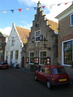 A traditional European building with a brick facade, multiple windows with shutters, and a red car parked in front.