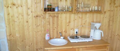 A wooden cabin interior with a kitchenette, including a sink, coffee maker, and shelves with jars and glasses.