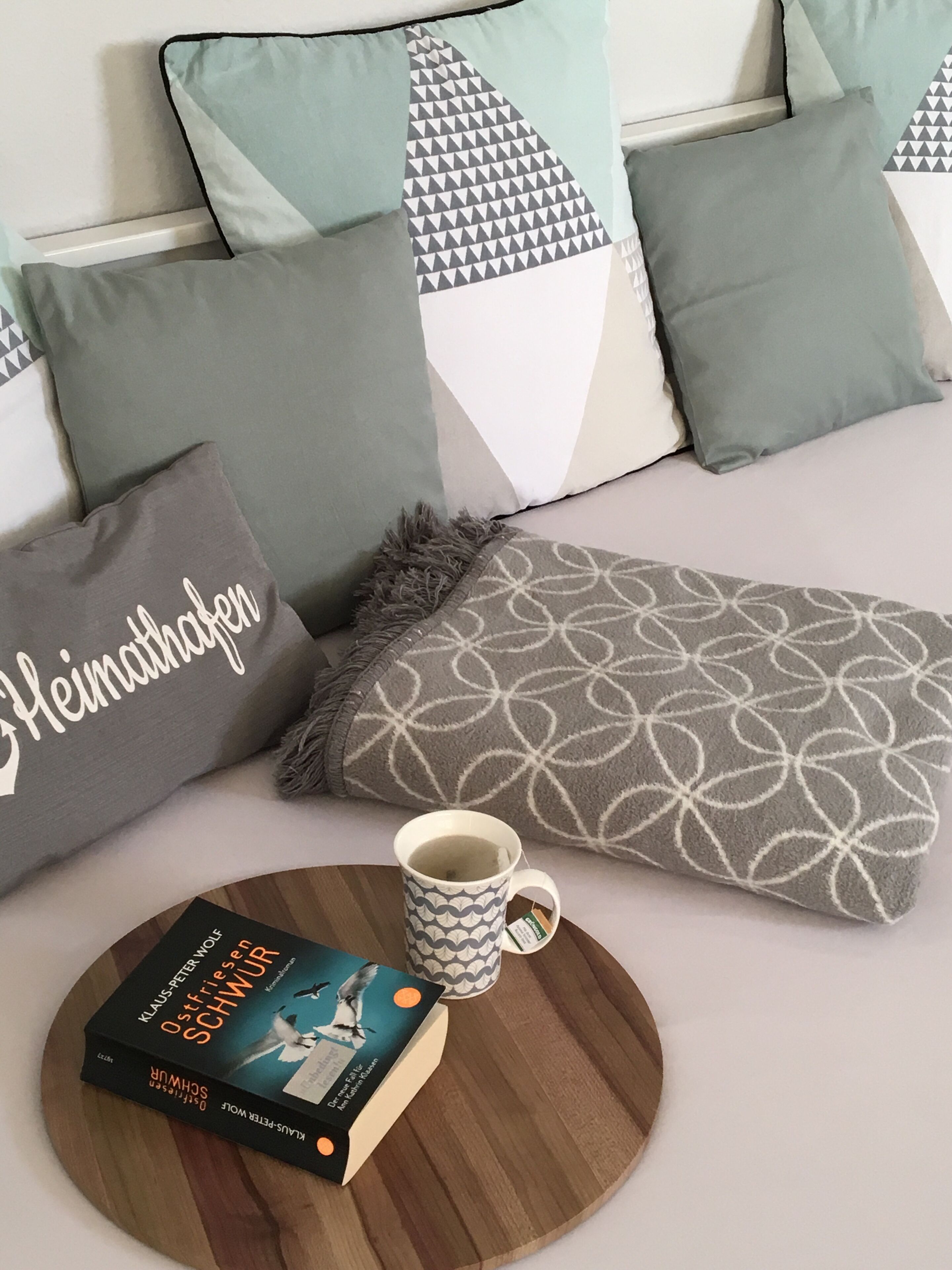 Un lit bien fait, agrémenté d’oreillers aux motifs géométriques, un livre intitulé « Schwüre » de Klaus Peter Wolf et une tasse posée sur un plateau en bois rond.