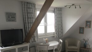 A room with a television on a dresser, a dining table with chairs, a window with curtains, and a wooden beam ceiling. - Last-Minute Summer Deals! A vacation right on the beach! (Neuharlingersiel)