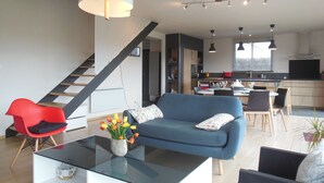 A modern living room with a blue sofa, a glass coffee table, and a red chair. There is a staircase leading to another level, a kitchen area with a dining table, and a large window providing natural light. - bright, contemporary new villa 2 minutes from beautiful beaches (Santec)