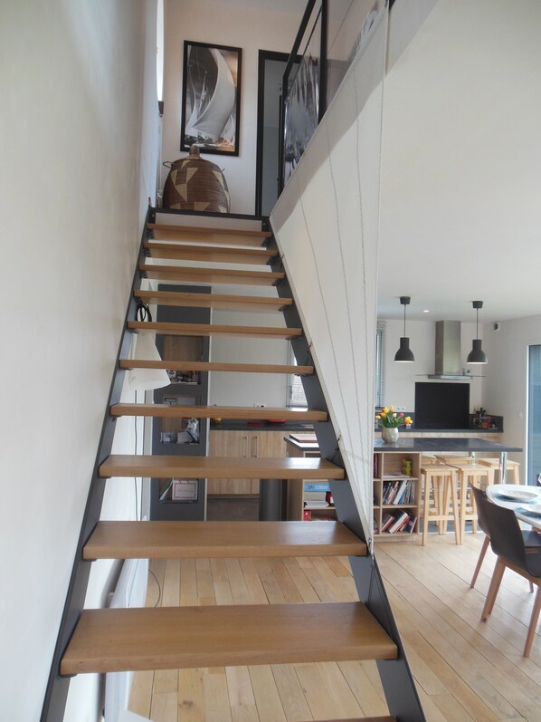 A modern staircase with wooden steps and metal railings, leading to a kitchen area with a dining table and chairs. - bright, contemporary new villa 2 minutes from beautiful beaches (Santec)