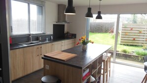 A modern kitchen with wooden cabinets, a central island with a cutting board, a sink, and a stove. There is a window with blinds, a hanging light fixture, and a view of a garden through the glass door. - bright, contemporary new villa 2 minutes from beautiful beaches (Santec)