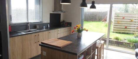 A modern kitchen with wooden cabinets, a central island with a cutting board, a sink, and a stove. There is a window with blinds, a hanging light fixture, and a view of a garden through the glass door.