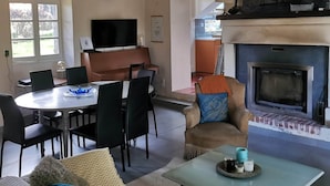 A cozy living room with a fireplace, a dining area with a round table, and a kitchen visible through an open doorway. - Nestled in a wooded park, with the beach just 3 minutes away (Pénestin)
