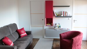 Un salon avec un canapé gris, des coussins rouges, une table basse blanche, un fauteuil rouge et une bibliothèque contenant des livres et des objets décoratifs.