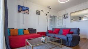 Una sala de estar moderna con un sofá rojo y azul, una mesa de centro de vidrio y una pared blanca decorada con obras de arte y un espejo.