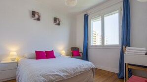 Un dormitorio con cama, mesitas de noche, una silla, una mesa con toallas y una ventana con cortinas.
