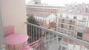 Un balcón con una mesa y sillas rosadas, con vistas a un paisaje urbano con edificios y cielo despejado.