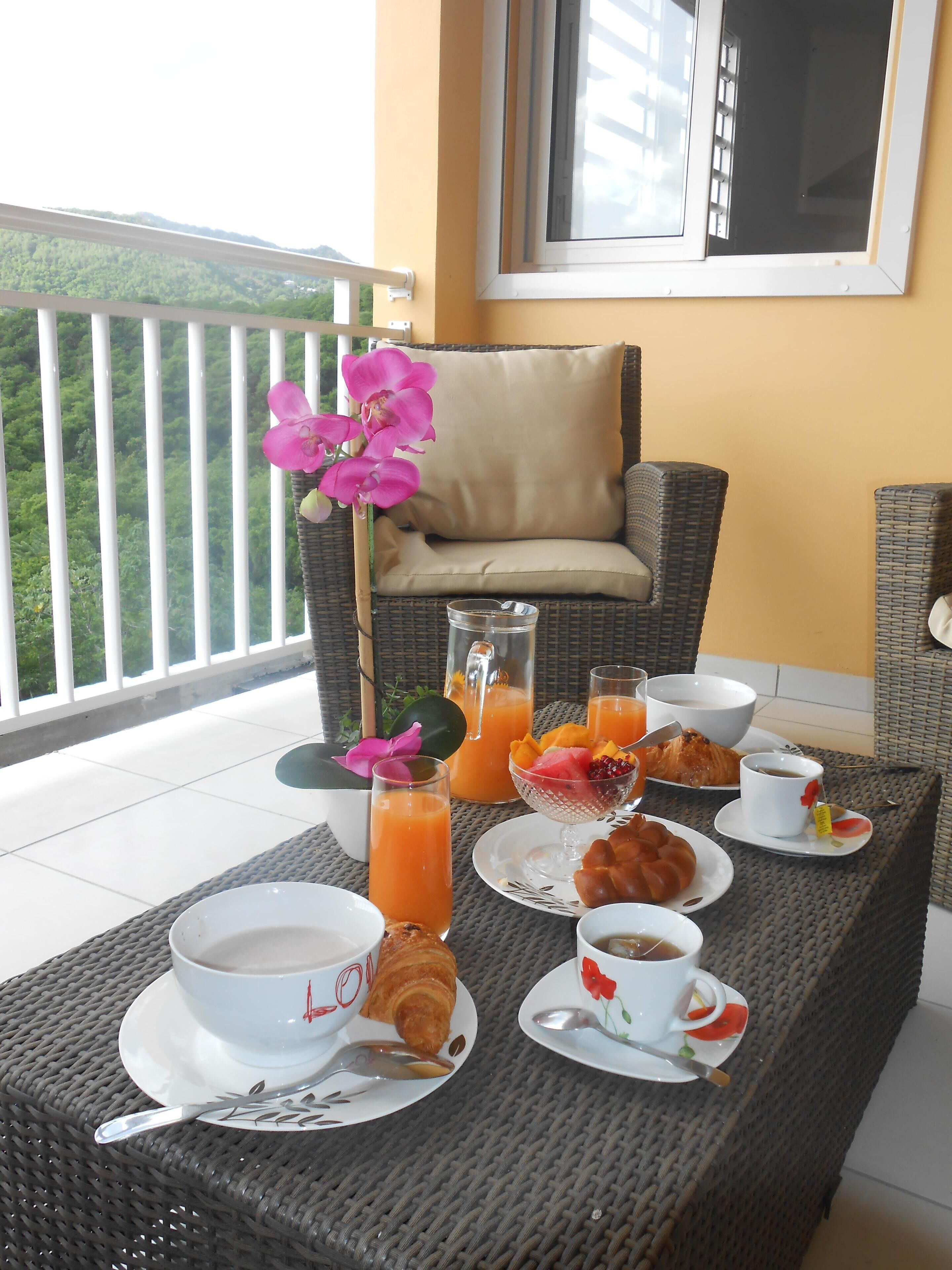 Ein Frühstücksbuffet auf einem Balkon mit Korbmöbeln, einem Tisch mit Tellern, Tassen und verschiedenen Speisen und Getränken.