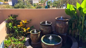 Une cour intérieure avec quatre grands pots rouillés remplis d’eau et agrémentés de jeux d’eau, entourée de différentes plantes et offrant une vue sur des bâtiments en arrière-plan.