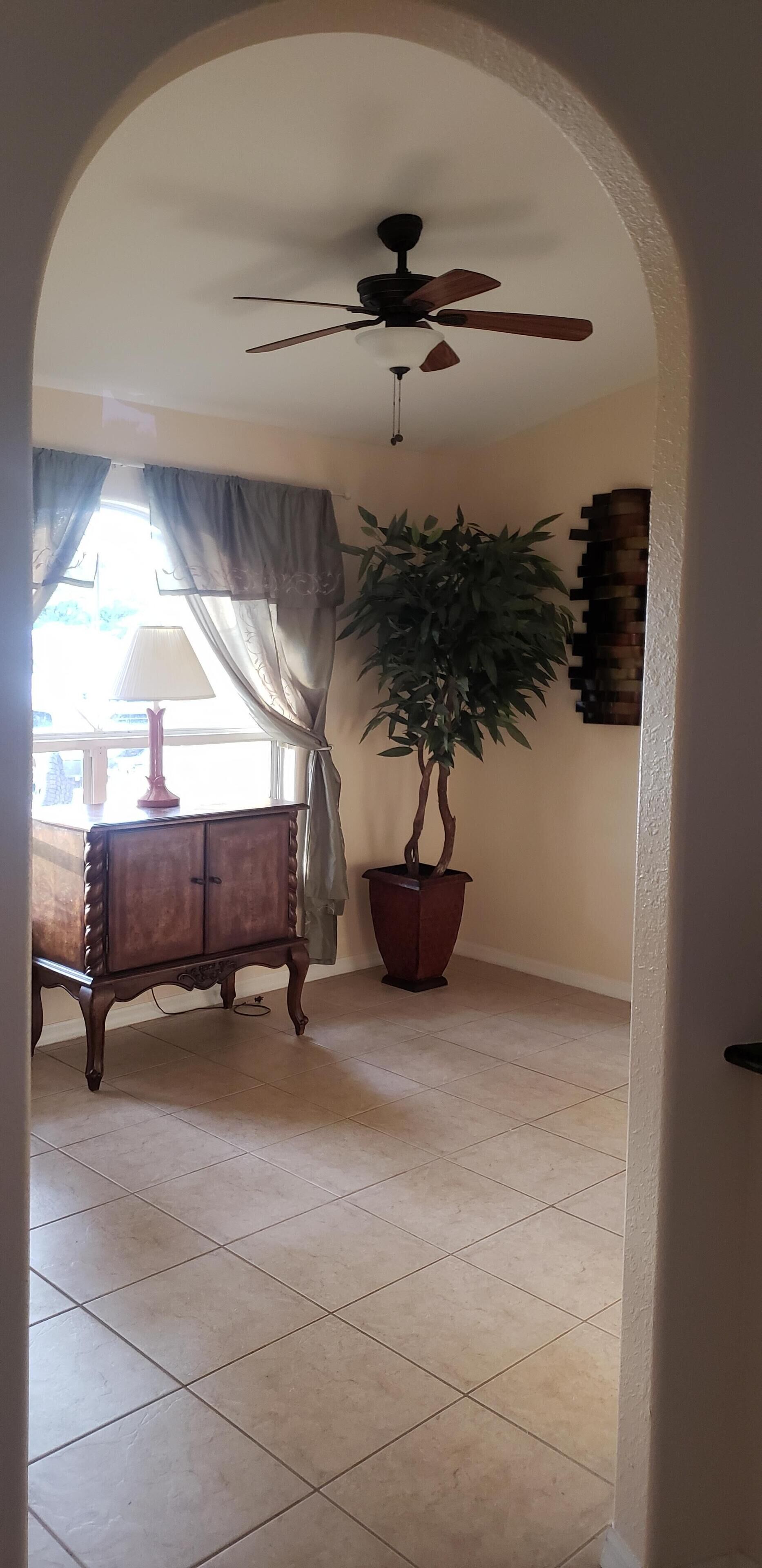 Un portail en arc mène à une pièce dotée d’un ventilateur de plafond, d’une plante en pot, d’une armoire en bois et d’une fenêtre avec des rideaux.