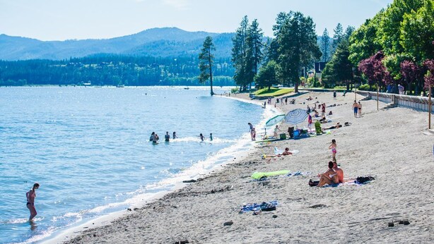 Une plage où des gens se prélassent au soleil et se baignent, entourée d’arbres et avec des montagnes en arrière-plan.