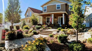 Une maison à deux étages avec un porche à l’avant, un drapeau et un jardin bien entretenu, agrémenté de diverses plantes et d’un chemin en pierre.