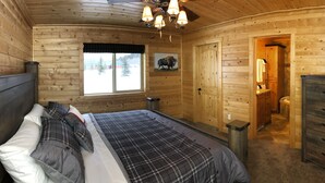 A wooden cabin bedroom with a bed, a dresser, a window with a view of trees, and a door leading to a bathroom.
