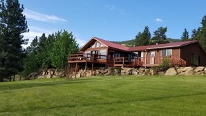 Une cabane en bois avec un toit rouge, une terrasse et des fondations en pierre, entourée d’un champ verdoyant et d’arbres.