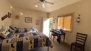 Une chambre avec deux lits, un bureau, une chaise, un ventilateur de plafond et une fenêtre avec des stores.
