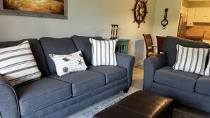 Una sala de estar con dos sofás grises, almohadas a rayas, un otoman marrón, un cuadro en la pared y un reloj.