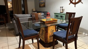 Un comedor con una mesa de vidrio, sillas de madera con cojines azules, un adorno con timón de barco y un cuadro en la pared.