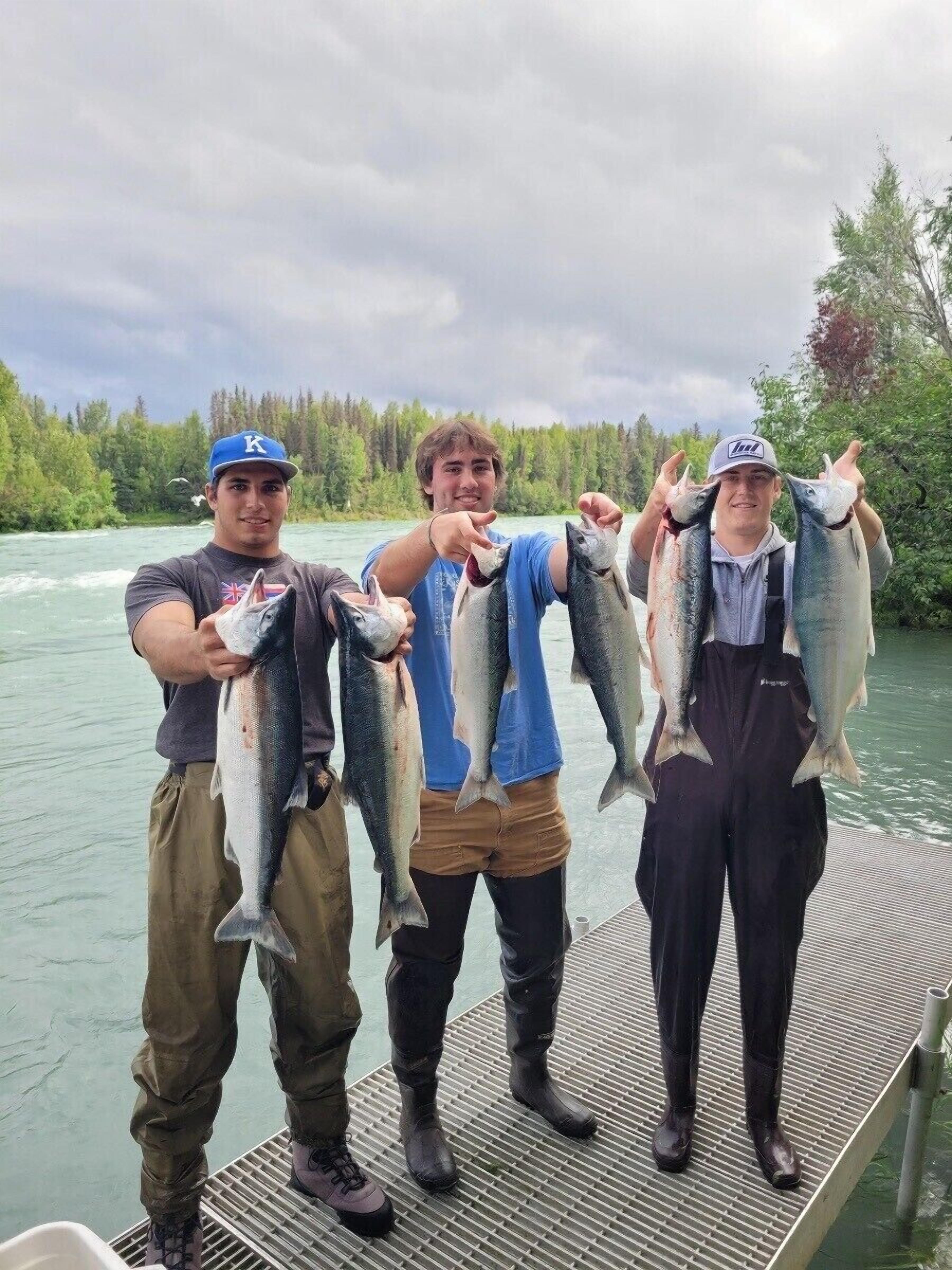 Trois personnes tenant des poissons sur un quai, avec une rivière et une forêt en arrière-plan.