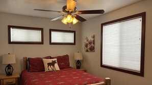 Ein Schlafzimmer mit einem hölzernen Bettgestell, einer roten Steppdecke mit Tiermotiven, zwei Nachttischlampen, einem Deckenventilator und drei Fenstern mit Jalousien.