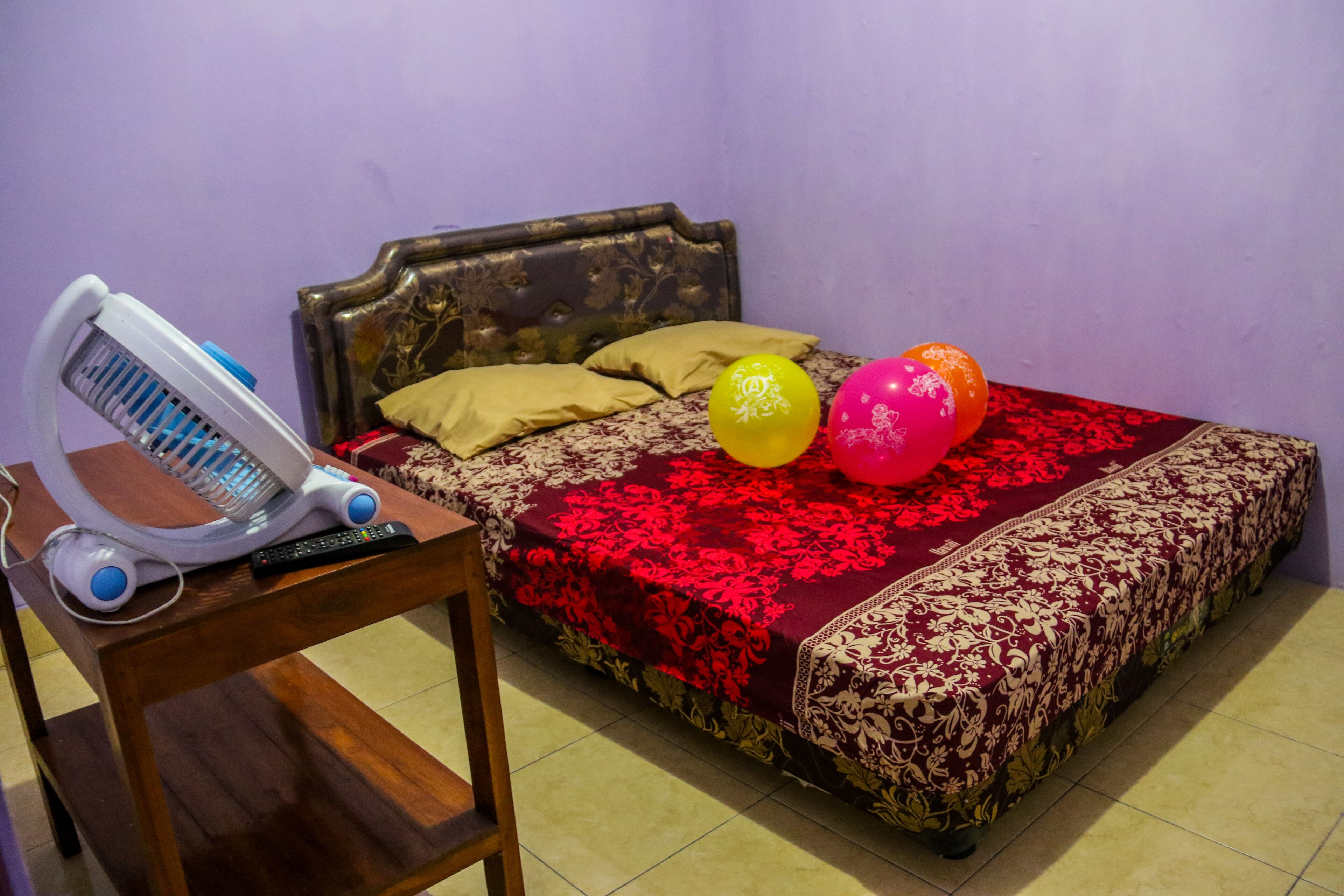 Un lit recouvert d’une couvre-lit aux motifs rouges et dorés, une table de chevet sur laquelle se trouve un radiateur électrique blanc, et deux ballons posés sur le lit.