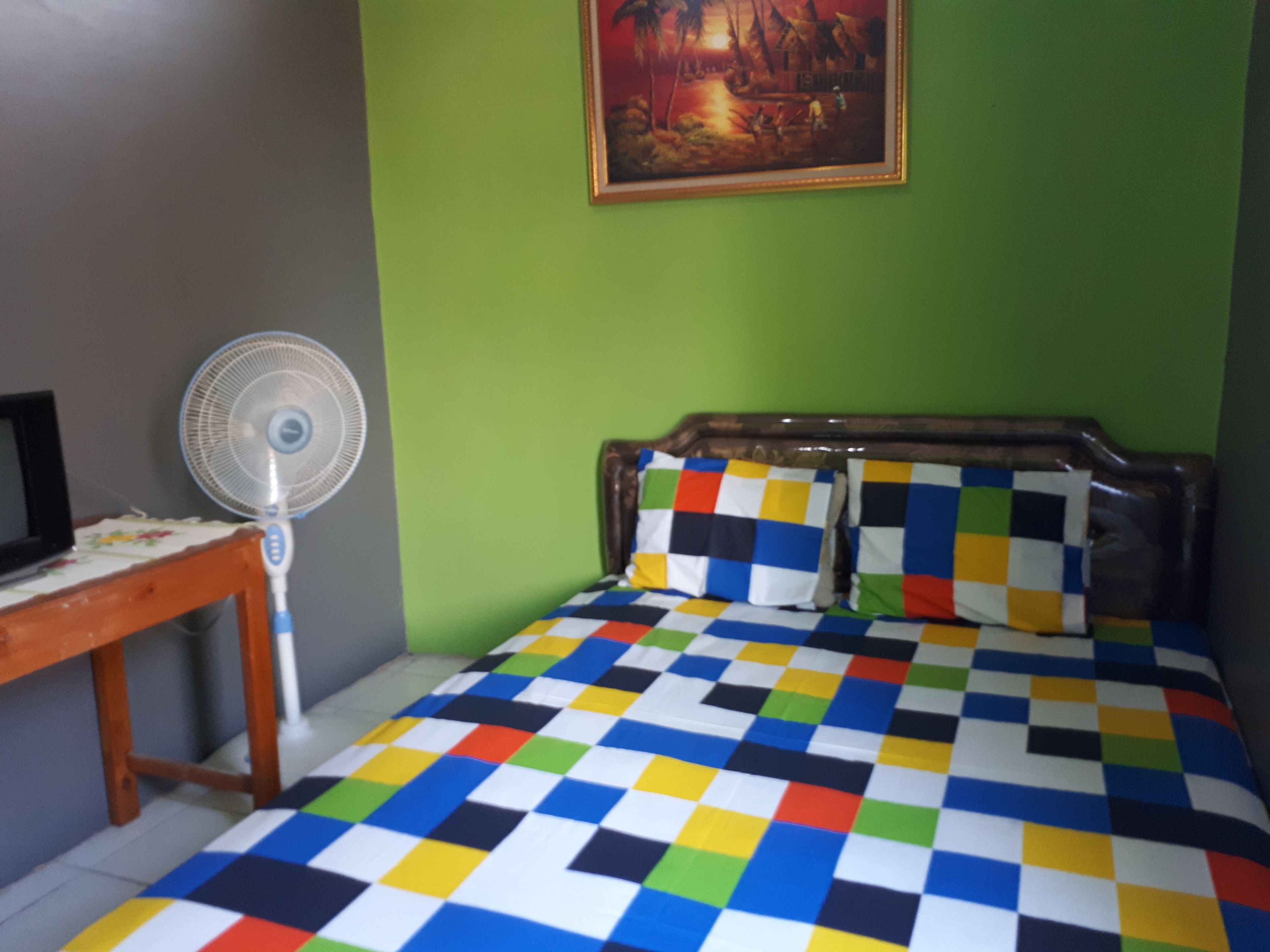 A room with a bed, a fan, a table, and a painting on the wall.